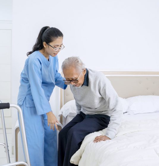 Beautiful nurse helping senior patient to sit on the bed while standing in the bedroom